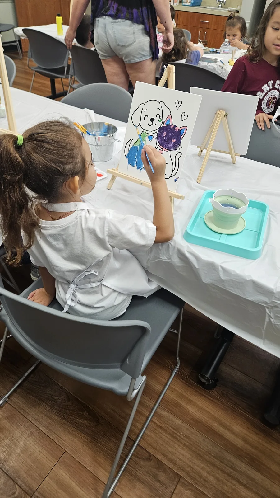 Child seen from behind painting a blue-and-purple puppy and kitten canvas on a small easel