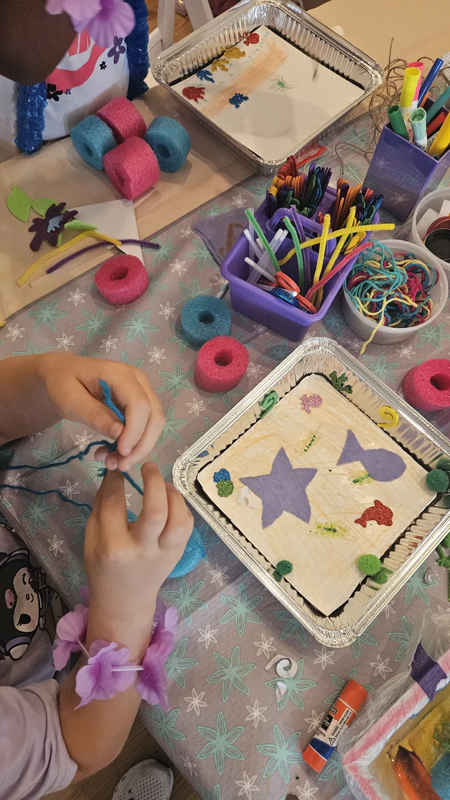 Close-up of hands threading yarn beside a decorated foil tray with felt shapes, pom-poms, and pipe cleaners