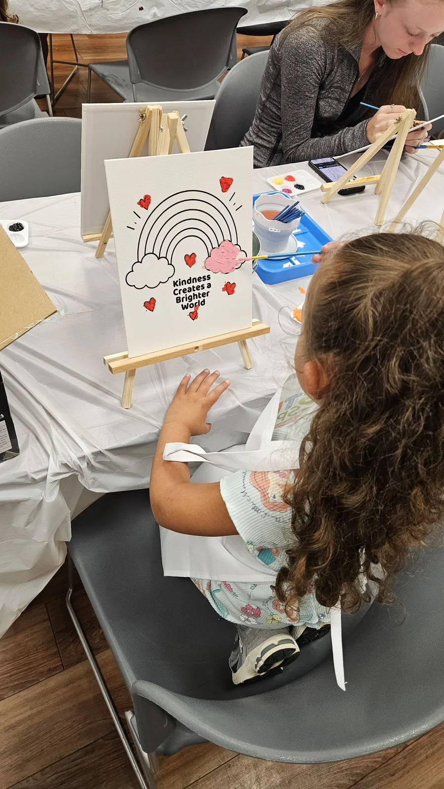 Child seen from behind adding pink paint to a rainbow-and-hearts canvas on a tabletop easel
