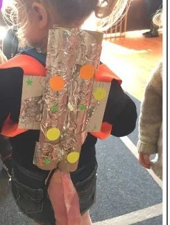 Child wearing a handmade cardboard jet pack covered in foil, colorful decorations, and fabric flames, photographed from behind